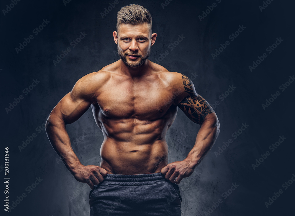 Obraz premium A handsome shirtless tattooed bodybuilder with stylish haircut and beard, wearing sports shorts, posing in a studio. Isolated on a dark background