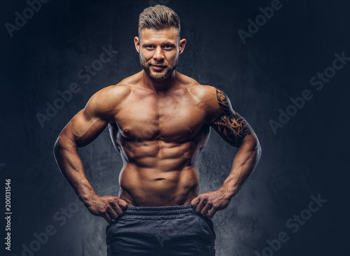 A handsome shirtless tattooed bodybuilder with stylish haircut and beard, wearing sports shorts, posing in a studio. Isolated on a dark background