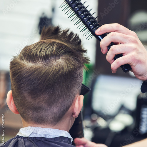 Barber is using hairdryer and hairbrush to make a styling to a Caucasian boy.