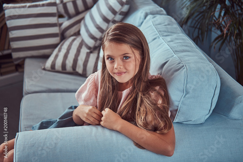 Portrait of a smiling little girl with long brown hair and piercing glance, lying on a sofa at home