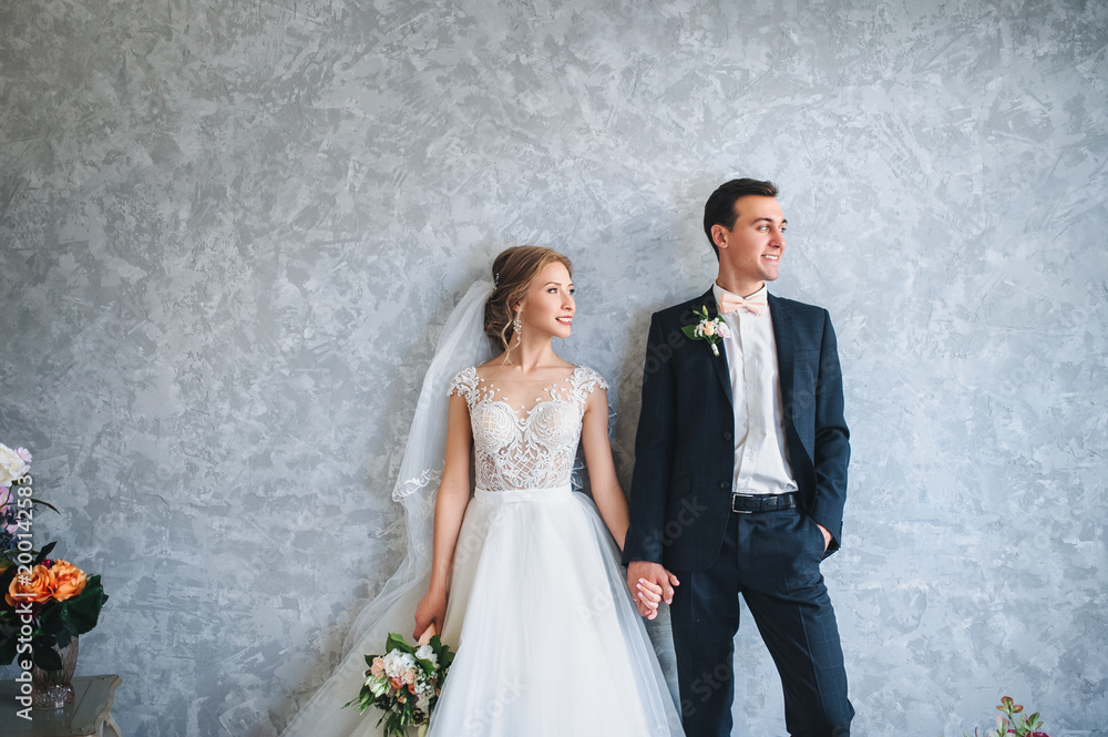 The young groom holds a beautiful bride in her hand in a lace dress near the gray wall, in a modern studio. Portrait of a newlywed couple.