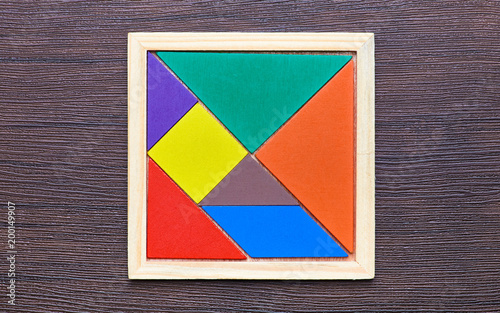 A tangram puzzle consisting of colored pieces with geometric shapes, collected in a square on a dark wooden background