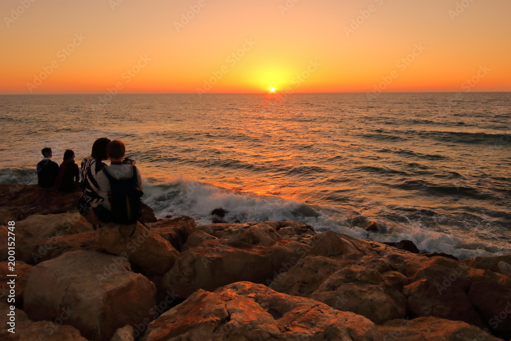 Romantyczny zachód słońca nad morzem, umocnione kamieniami nabrzeże i woda oświetlona złotymi promieniami zachodzącego słońca, przytulona para siedzi tyłem,  na kamieniach, wpatruje się w słońce