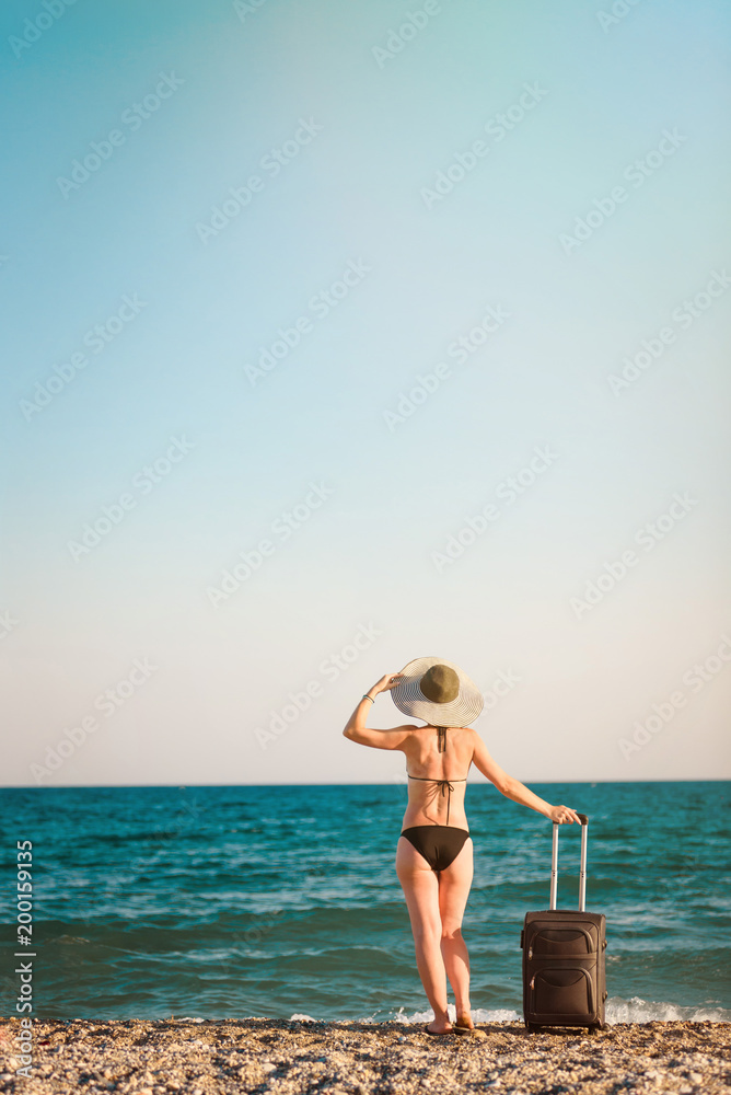 Young beautiful woman in swimsuit and hat with large suitcase on tropical beach, wyth sea on background Young beautiful woman in swimsuit and hat with large suitcase on tropical beach, wyth sea on background