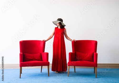 Beautiful young woman in red dress and red chair, in white room with blue floor