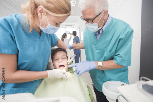Boy with perfect teeth at the dentist doing check up with two doctors while other doctors analyze x-ray in the background - oral hygiene health care concept
