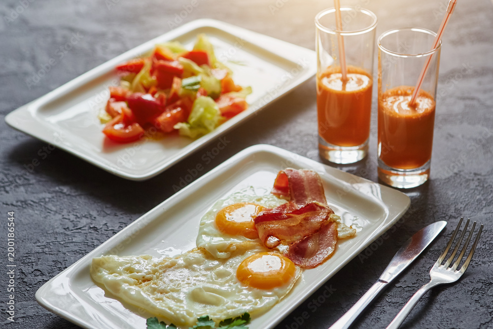 Breakfast from fried egg, bacon, vegetable salad and carrot juice. On a dark stone background Breakfast from fried egg, bacon, vegetable salad and carrot juice. On a dark stone background
