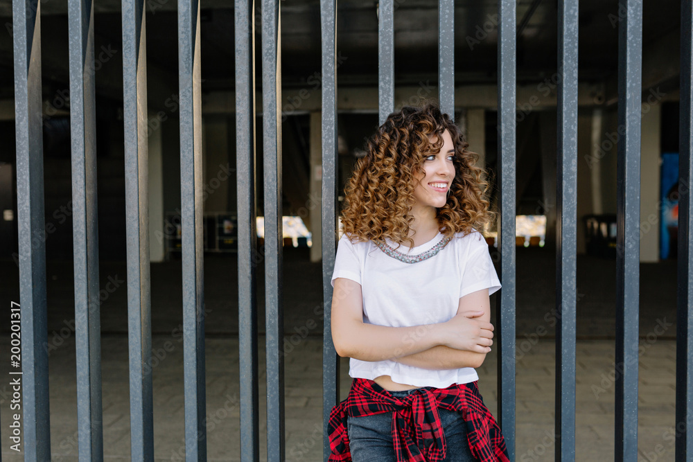 image of fantastic girl with open smile and curly style hair dressed in white shirt and checkered shirt in the city. She is looking aside. Real happy emotions. Place for text.