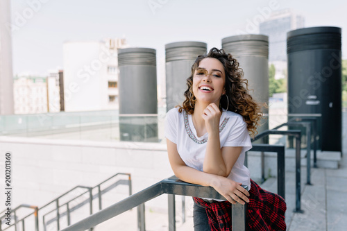Obraz na płótnie image of stylish beautiful girl with open smile and curly style hair dressed in white shirt in the city. She is laughing at the camera. Real happy emotions. summer sunny day. w salonie