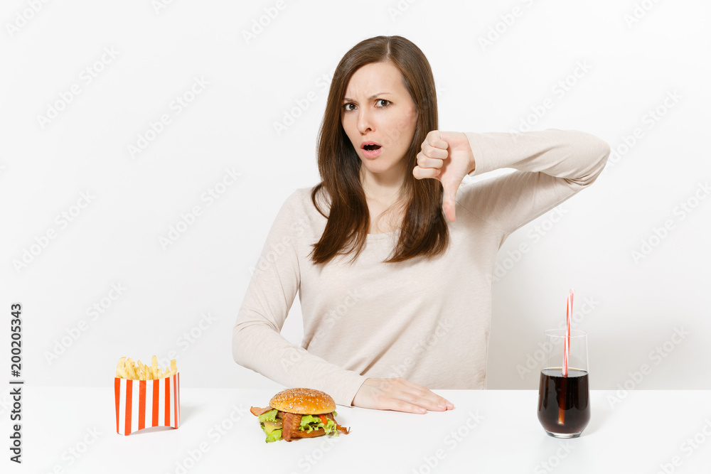 Irritated woman shows thumbs down at table with burger french fries cola in glass bottle isolated on white background. Proper nutrition or American classic fast food. Advertising area with copy space. Irritated woman shows thumbs down at table with burger french fries cola in glass bottle isolated on white background. Proper nutrition or American classic fast food. Advertising area with copy space.