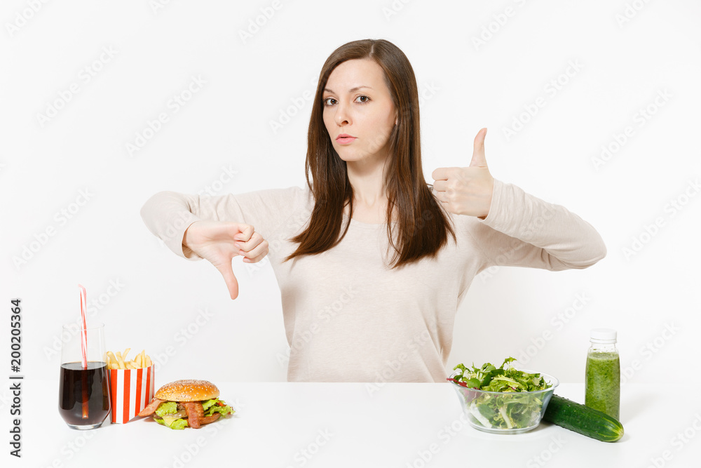Woman shows thumbs up down with green detox smoothies, salad in glass bowl, cucumber burger cola in bottle isolated on white background. Proper nutrition, healthy lifestyle, fast food, choice concept. Woman shows thumbs up down with green detox smoothies, salad in glass bowl, cucumber burger cola in bottle isolated on white background. Proper nutrition, healthy lifestyle, fast food, choice concept.