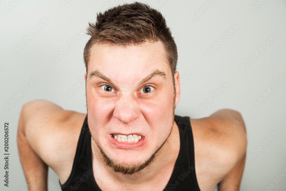 Fototapeta premium Young funny bearded guy dressed in black tank top sitting on the toilet on gray background.