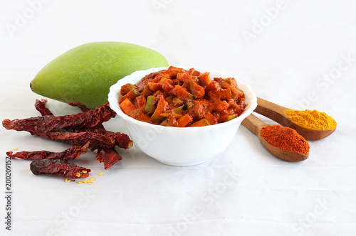 Pickles, an Indian popular side dish made from a type of mango known as totapuri, in a bowl, and in the background are the fruit, red chilies and chili and turmeric powder.