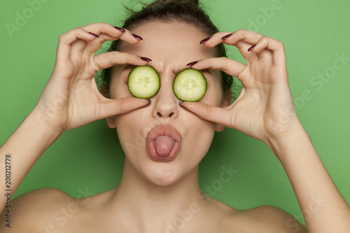 Happy sexy woman posing with slices of cucumber on her face on green background