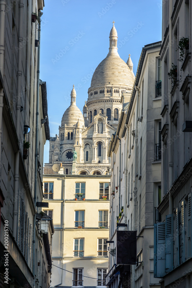 The Basilica of the Sacred Heart of Paris seen through a narrow street between old parisian buildings under a clear blue sky.