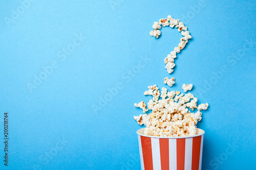 Concept of which film to choose. Spilled popcorn in the form of question mark and a paper bucket in red strip on blue background. Copy space for text