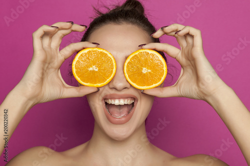 Happy young woman posing with slices of oranges on her face on pink background