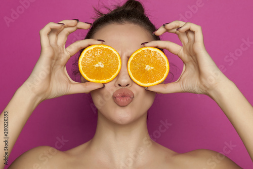 Young sexy woman posing with slices of oranges on her face on pink background
