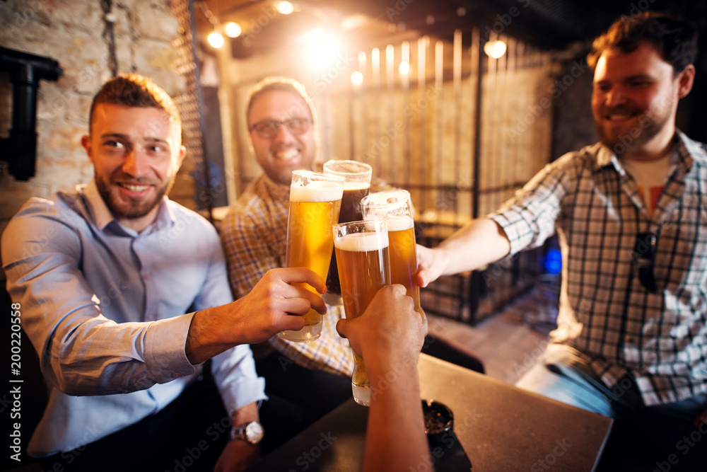 A group of young cheerful men clinking glasses with a beer in the sunny pub after work. A group of young cheerful men clinking glasses with a beer in the sunny pub after work.