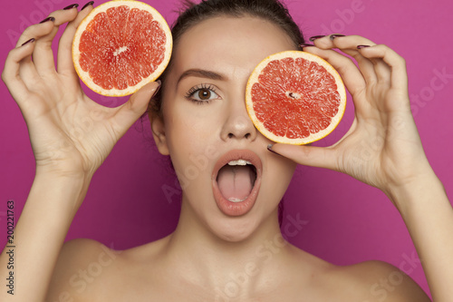 Young surprised woman posing with slices of red grapefruit on her face on pink background