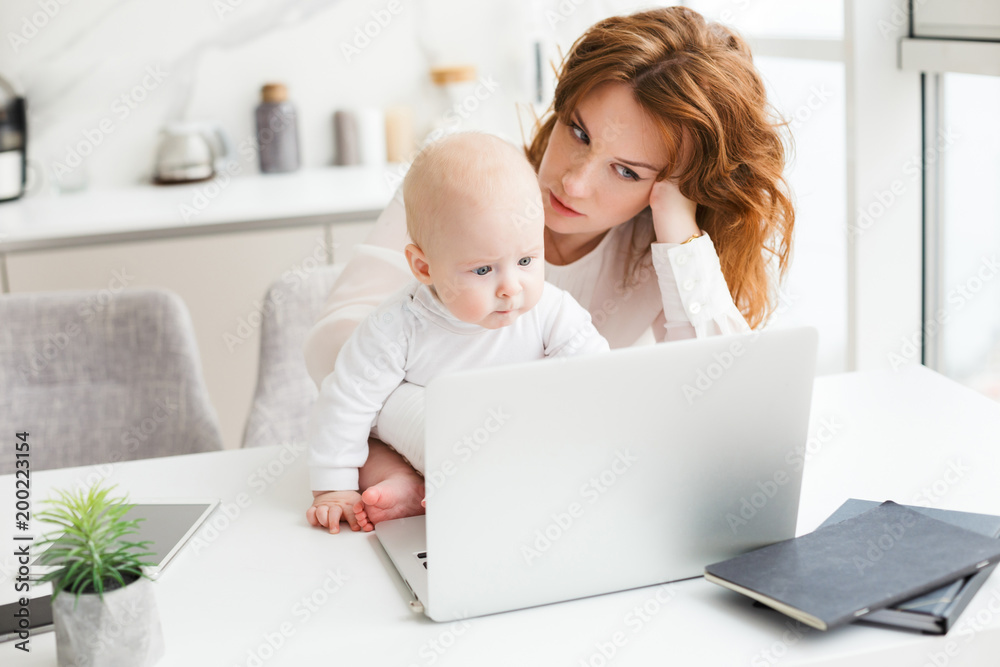 Portrait of tired business woman sitting at the table and working on laptop while holding her little baby near isolated Portrait of tired business woman sitting at the table and working on laptop while holding her little baby near isolated