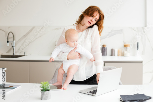 Portrait of business woman standing with her little baby in hand and talking on her cellphone near table with laptop isolated.