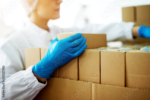Close up of young focused female worker in sterile cloths is folding boxes on stacks of boxes in factory storage room.