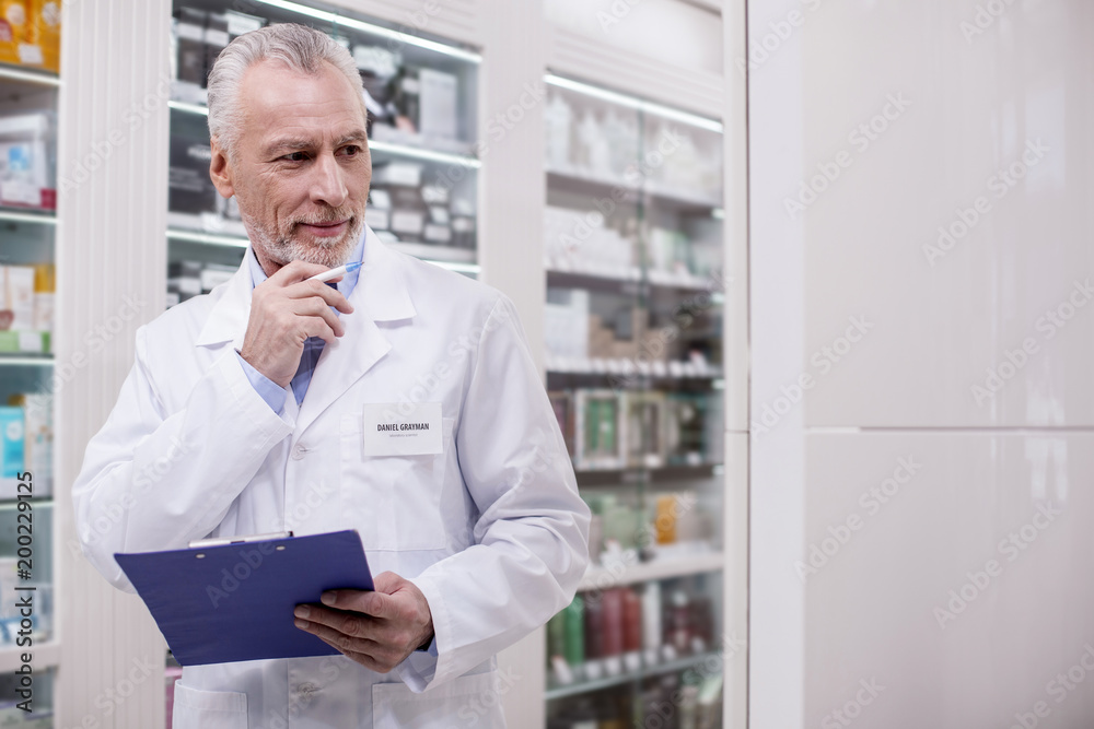 Responsible work. Senior attractive male pharmacist using clipboard while touching face