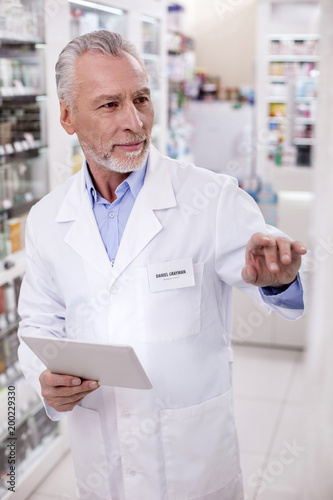 Online registration. Mature pensive male pharmacist holding tablet while rising hand