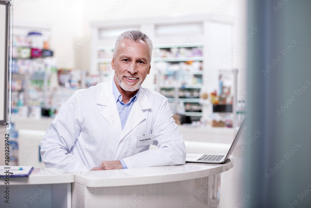 Working day. Mature gay male pharmacist looking at camera while leaning on stand Working day. Mature gay male pharmacist looking at camera while leaning on stand