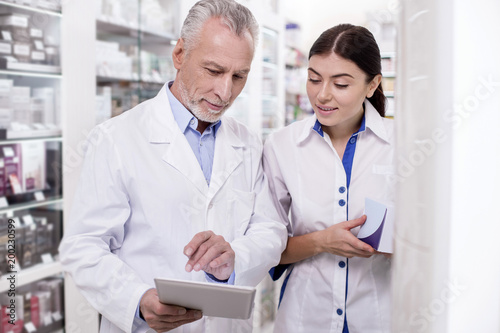 Valuable advice. Pleasant two pharmacists gazing at tablet screen while standing in drugstore