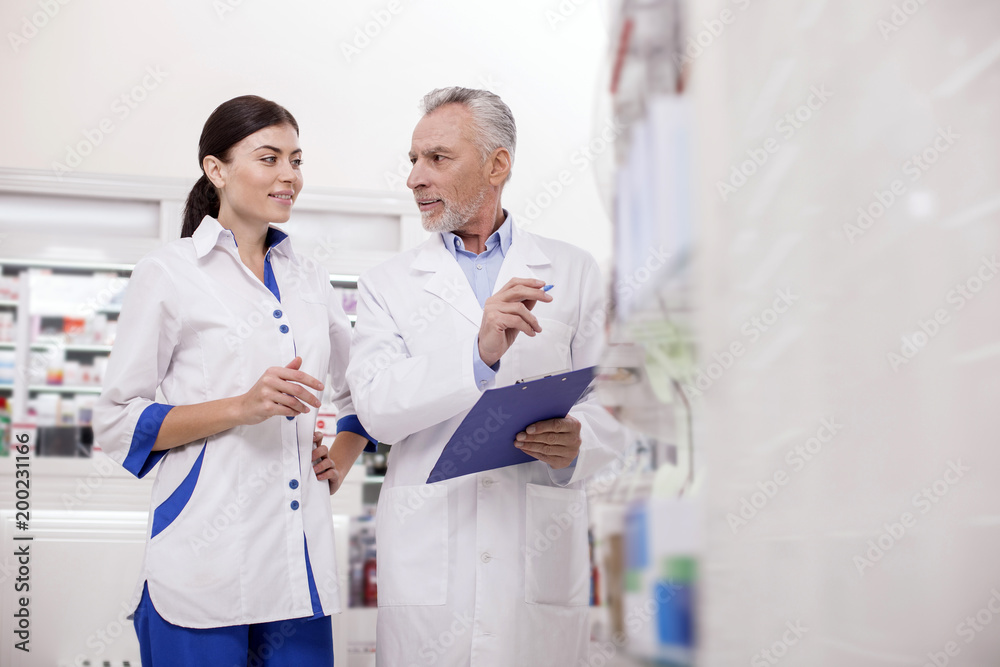 Obraz premium New information. Low angle of successful pleasant two pharmacists chatting while supervising products