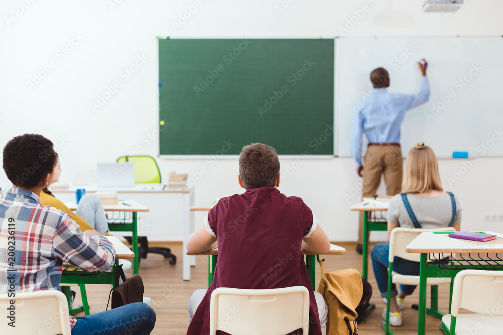 Rear view of schoolchildren and teacher writing on white board