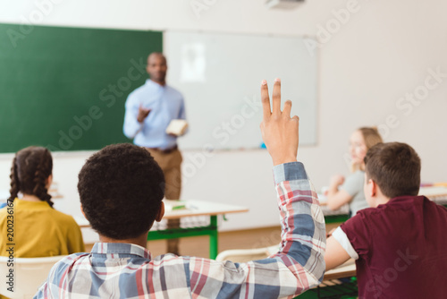 Rear view of african american boy with arm up in classroom with teacher and classmates