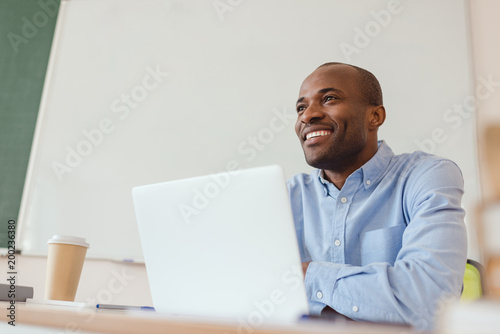Low angle view of smiling african american teacher sitting at table with laptop and coffee cup