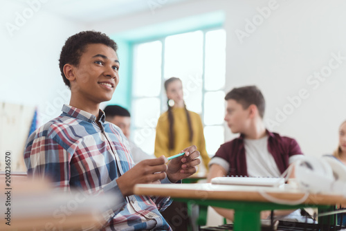 Low angle view of african american high school student holding pencil in hand and classmates sitting behind