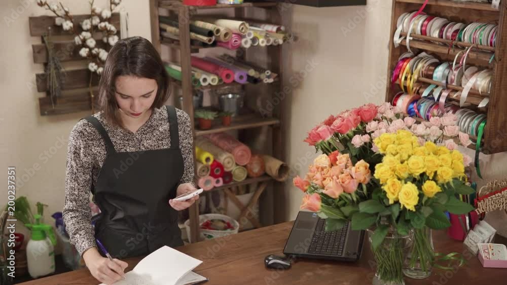 Shot from above of a attractive brunette woman working in a flower shop. She is holding phone and writing in a diary. Shot from above of a attractive brunette woman working in a flower shop. She is holding phone and writing in a diary.