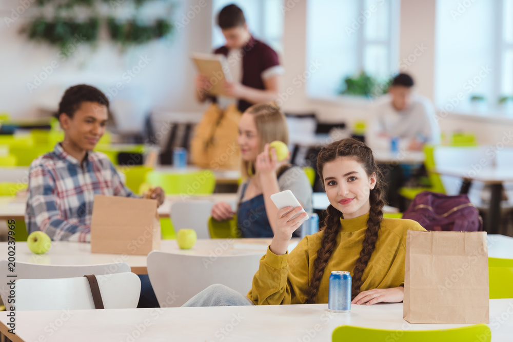 Obraz premium smiling teen schoolgirl sitting at school cafeteria with classmates and looking at camera