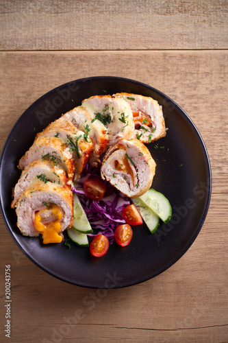 Chicken wrapped with ham and cheddar cheese; vegetables in black plate on wooden table. vertical