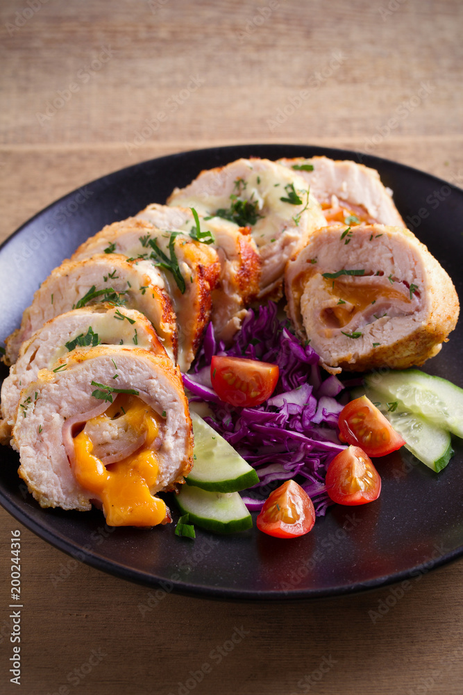 Chicken wrapped with ham and cheddar cheese; vegetables in black plate on wooden table. vertical Chicken wrapped with ham and cheddar cheese; vegetables in black plate on wooden table. vertical
