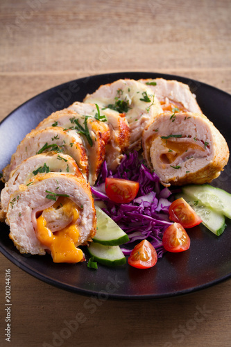 Chicken wrapped with ham and cheddar cheese; vegetables in black plate on wooden table. vertical