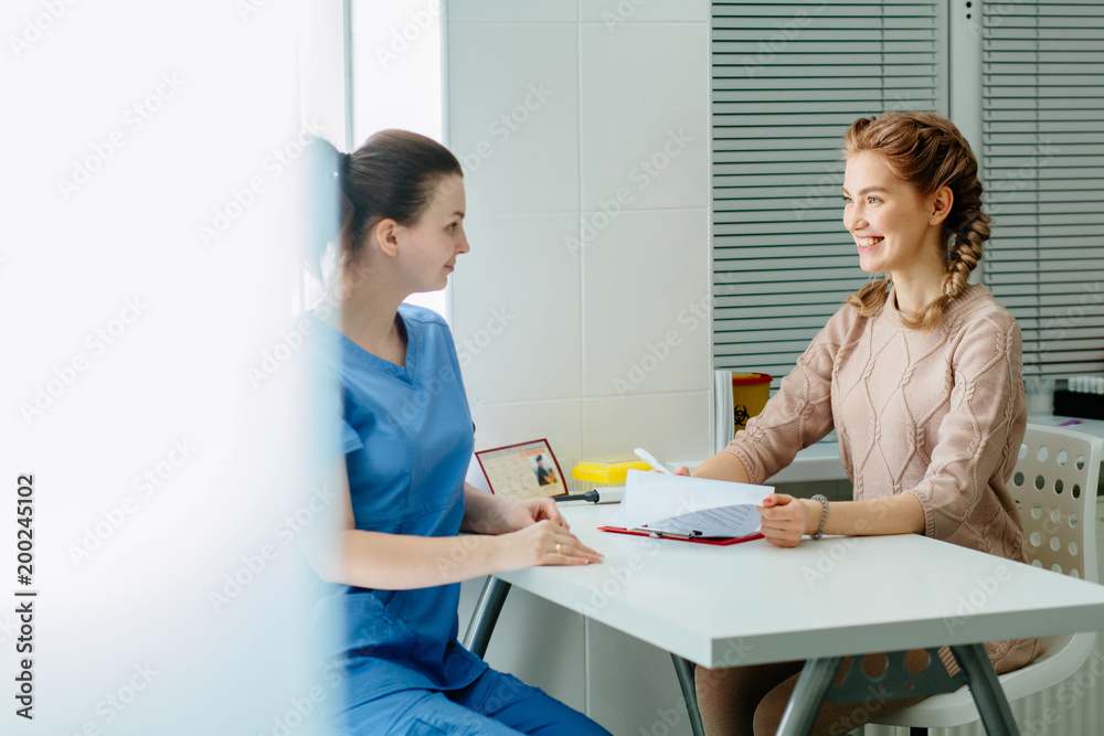 Beautyful blonde woman sitting in cabinet and talking to doctor about signing paper. Women's health and medicine concept. Beautyful blonde woman sitting in cabinet and talking to doctor about signing paper. Women's health and medicine concept.