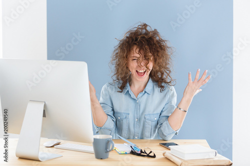 Happy positive young successful business woman screaming yes with clenched fists, has overjoyed expression, celebrating success, smiling and looking at the camera while working on computer in office