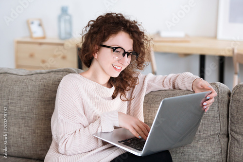 Portrait of attractive yound red hair caucasian businesswoman in eyeglasses at home working on laptop computer remotely. Woman sitting on her couch using computer,uses wireless internet connection