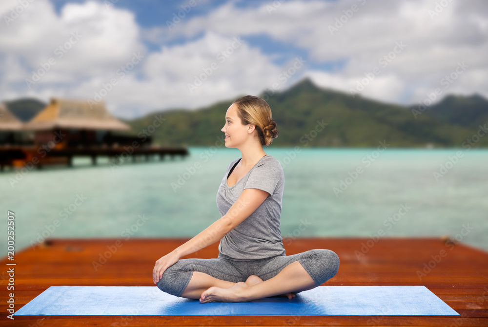 fitness, sport, people and healthy lifestyle concept - woman making yoga in twist pose on wooden pier over island beach and bungalow background