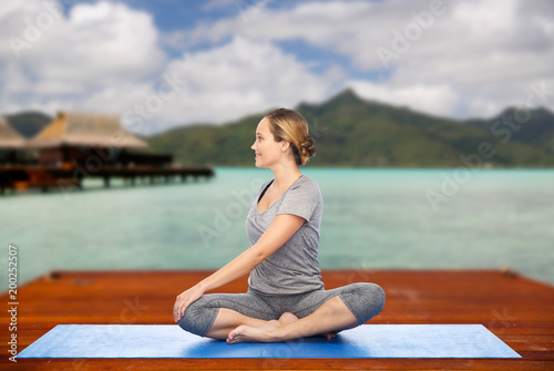 fitness  sport  people and healthy lifestyle concept - woman making yoga in twist pose on wooden pier over island beach and bungalow background