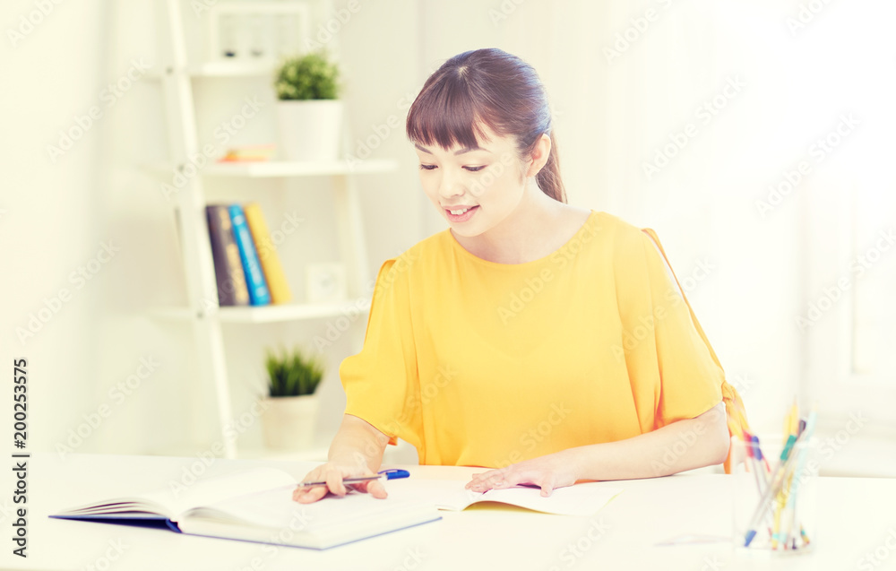 people, education, high school and learning concept - happy asian young woman student with book and notepad writing at home people, education, high school and learning concept - happy asian young woman student with book and notepad writing at home