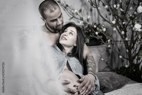 Black and white photo of smiling international couple of man with bare chest hugging his brunette pregnant wife while sitting on gray cozy bed