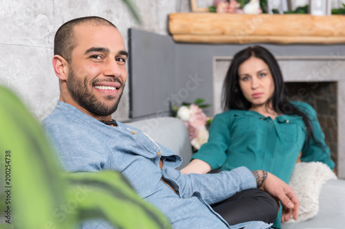 Smiling international couple of man with beard and his brunette wife sitting on the sofa