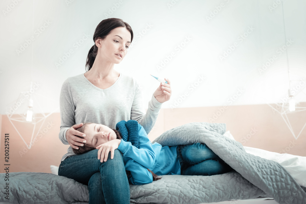 You need a rest. Occupied caring pleasant woman sitting on the bed holding the thermometer touching to her daughter.